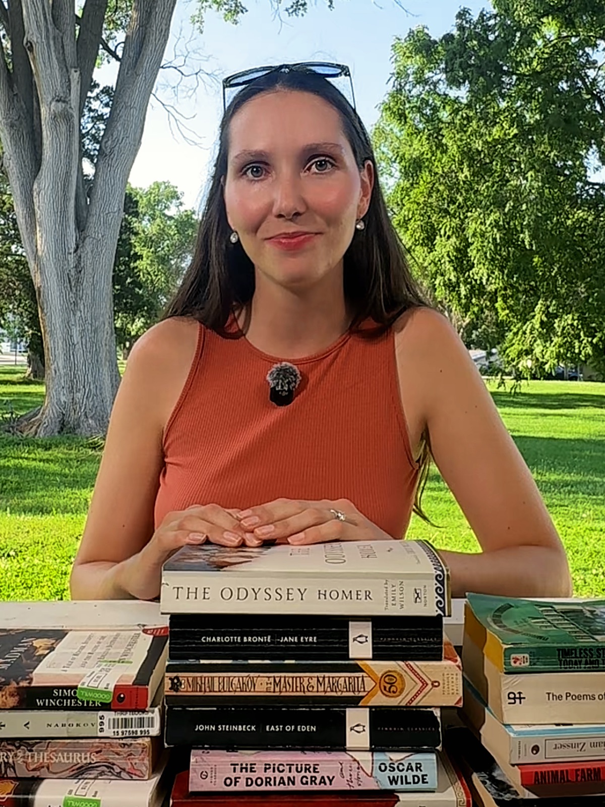 Katherine outdoors behind a stack of classic books — The Odyssey, Jane Eyre, The Master and Margarita, East of Eden, and more